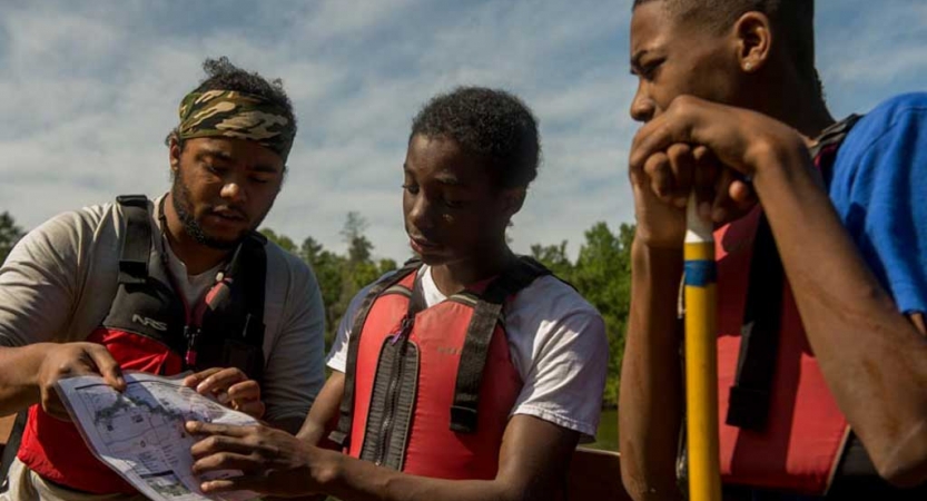 Three people wearing life jackets examine a map. One of them is leaning on a paddle. 